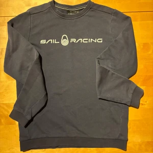 Mörkblå Sail Racing sweatshirt - Mörkblå sweatshirt från Sail Racing med vit logga framtill. Klassisk rund halsringning och långa ärmar med ribbade muddar. Skön och stilren tröja i bomull, perfekt för en chill och sportig look.