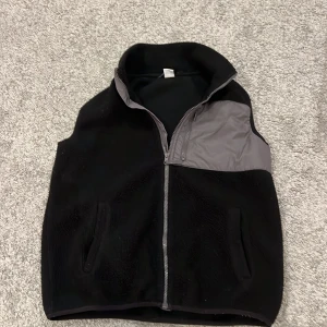 Svart fleeceväst med grå detalj - Svart fleeceväst med hög krage och grå panel upptill på framsidan. Västen har dragkedja och två fickor framtill. Perfekt för lager-på-lager och chill dagar.