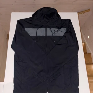 Svart vindjacka från Under Armour  - Säljer en svart vindjacka från Under Armour i storlek M. Jackan har en grå panel över bröstet med logga, huva med dragsko och hel dragkedja framtill. Tillverkad i lätt polyester och har en loose passform, perfekt för träning eller chill dagar.