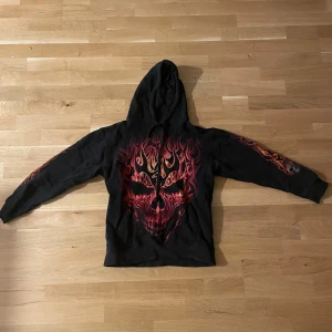 Svart hoodie med eldskalletryck - Svart hoodie med stora, detaljerade tryck av eldsflammor och dödskallar både fram och bak. Huvan har snörning och ärmarna har också eldiga detaljer. Perfekt för dig som gillar en edgy och unik stil. Nypris 699kr, använd 2 gånger 