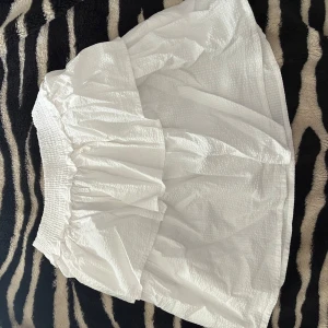 Vit volangkjol från Polo Ralph Lauren - Superfin vit kortkjol från Polo Ralph Lauren med volang och smockad midja. Kjolen har en luftig, lätt struktur som ger en fräsch och somrig känsla. Perfekt för dig som gillar en enkel men ändå söt stil.