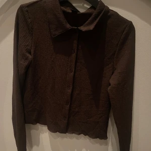 Brun skjorta från SHEIN med struktur - Mörkbrun skjorta från SHEIN i storlek L. Skjortan har en strukturerad, lätt skrynklig yta, klassisk krage och knäppning framtill. Långärmad och med lätt croppad passform. Perfekt för dig som gillar stilrena och enkla plagg med en twist.