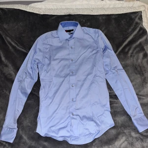 Ljusblå skjorta med knappar - Snygg ljusblå skjorta från Eterna med klassisk krage och långa ärmar. Skjortan har en slim passform och är tillverkad i mjuk bomull. Perfekt för dig som gillar stilrena och enkla plagg. Knappar hela vägen framtill. Skrynklig på bild då den legat i garderob länge.