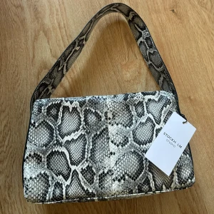 Axelväska i ormmönster från Stockh LM - Snygg axelväska från Stockh LM Studio med trendigt ormmönster i svart, grått och vitt. Väskan har en bred axelrem och rektangulär form, perfekt för att lyfta vilken outfit som helst. Materialet är syntet med struktur som påminner om ormskinn.