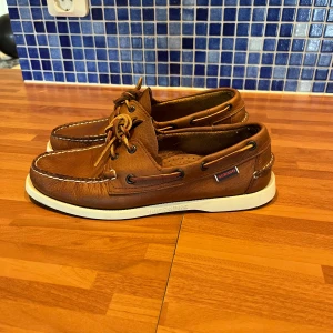 Bruna seglarskor från Sebago - Säljer ett par klassiska bruna seglarskor från Sebago med vit sula och snygga detaljer i skinn. Skorna har snörning runtom och kontrastsömmar framtill. Perfekt för dig som gillar tidlös stil och vill ha något som funkar till jeans eller shorts.