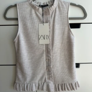 Beige ärmlös topp med volang från Zara - Supersöt beige ärmlös topp från Zara med knappar framtill och volangdetalj längs nederkanten. Toppen är ribbad och har en rund halsringning, perfekt för stilren look. 