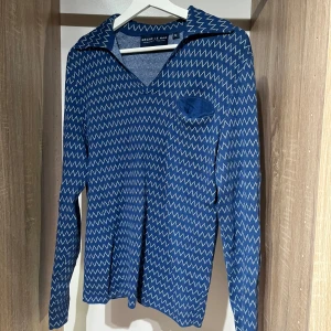 Blå mönstrad skjorta Grand Le Mar - Blå långärmad piké från Grand Le mar med chevronmönster. Skjortan har en öppen krage och en bröstficka med matchande tyg. 