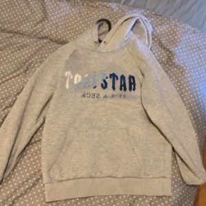 Grå hoodie från Trapstar med tryck - Säljer en grå hoodie från Trapstar med blått och vitt tryck på bröstet. Tröjan har huva med snörning och en stor magficka. Perfekt för dig som gillar streetwear och vill ha en bekväm och snygg look.