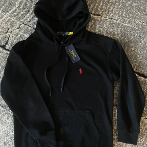 Svart hoodie från Polo Ralph Lauren - Svart hoodie från Polo Ralph Lauren med klassisk röd broderad logga på bröstet. Tröjan har huva med snörning och en stor magficka. Tillverkad i mjuk bomull, perfekt för hösten och mysigt material!                                                                   Nypris: ca 2200kr. Mitt pris: 950kr 