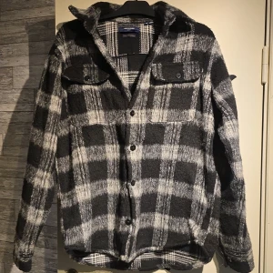 Rutig overshirt från Jack & Jones - Snygg overshirt från Jack & Jones i svart och grått rutigt mönster. Modellen har två bröstfickor med knappar, klassisk krage och långa ärmar med knappslå. Materialet är mjukt och lite fluffigt, perfekt för lager-på-lager. Passar dig som gillar street och chill stil.