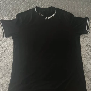Svart t-shirt med tryck på kragen XL - Svart t-shirt i storlek XL med vit text 'Improve Aesthetic' tryckt runt kragen. T-shirten har korta ärmar med vita ränder på ärmsluten och en klassisk rak passform. Perfekt för dig som gillar streetwear och stilrena detaljer.