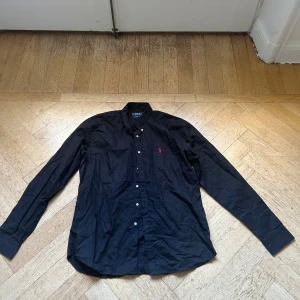 Svart skjorta från Polo Ralph Lauren - Svart skjorta från Polo Ralph Lauren med klassisk krage och knäppning framtill. Skjortan har lång ärm och den ikoniska röda logotypen broderad på bröstet. Tillverkad i bomull för en skön känsla och snygg passform.