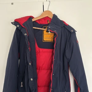 Mörkblå pufferjacka från A One - Säljer en mörkblå pufferjacka från A One med röd insida och detaljer. Jackan har huva med riktig kanin päls, dragkedja och tryckknappar framtill samt praktiska fickor. Perfekt för kalla dagar och designad av Bernadotte & Kylberg.  Köpt för 1900 med riktig kanin päls och perfekta vinter jackan!