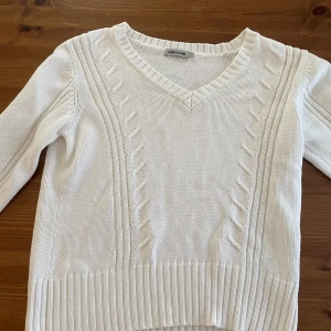 Vit stickad tröja från Long Island - Vit stickad tröja från Long Island med v-ringning och snygga kabelstickade detaljer framtill. Tröjan har långa ärmar och bred ribbad mudd vid ärmslut och nederkant. Perfekt för lager-på-lager och chill stil.