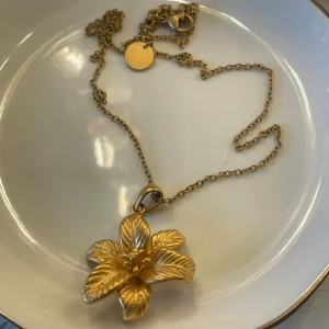 Guldigt blom-halsband från Olympia - Supersnyggt halsband i guldton från Olympia rings med ett stort, detaljerat blomformat hänge. Kedjan är tunn och har en rund bricka med märkets logga. Perfekt för dig som vill sticka ut med en unik accessoar med blommig vibe.