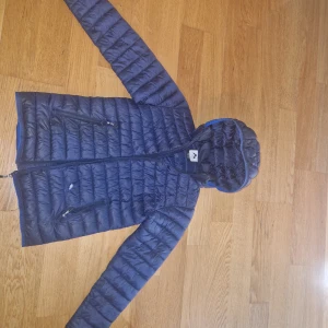 Blå McKinley pufferjacka med huva - Säljer en marinblå pufferjacka från McKinley med huva och två dragkedjeförsedda fickor. Jackan är lätt vadderad, har horisontella sömmar och reflekterande detalj bak. Perfekt för kyliga dagar och enkel att bära med sig. Litet hål på framsidan tyvärr. Säljes billigt