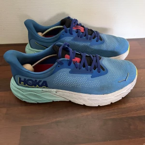 Blå Hoka Arahi 7 löparskor - Säljer ett par Hoka Arahi 7 löparskor i blått med turkos mellansula och gula detaljer. Skorna har snörning, ventilerande mesh-ovandel och en riktigt skön, tjock sula för extra dämpning. Perfekta för dig som gillar att springa långt och vill ha stabilitet och komfort.