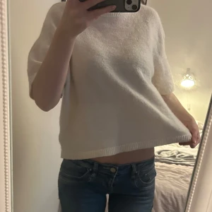 Vit stickad topp med kort ärm - Mysig vit stickad topp med korta ärmar och rund hals. Modellen är loose fit och har ribbad kant nedtill. Perfekt för dig som gillar enkel och clean stil. Passar till jeans eller kjol för en chill look.