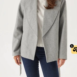 Grå omlott kappa från ONLY - Snygg grå omlott kappa från ONLY med bred krage och bälte i midjan. Jackan har lång ärm och rak passform, perfekt för dig som gillar stilren och enkel design. Materialet är mjukt och känns lyxigt mot huden. Passar dig som vill ha en chill men ändå classy look.