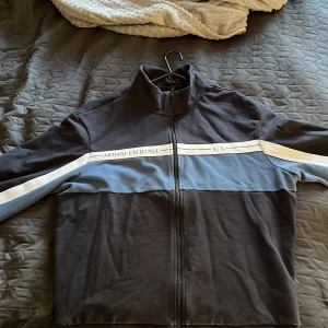 Svart och blå overshirt från Armani Exchange - Svart och blå overshirt från Armani Exchange med vit rand längs bröstet och ärmarna. Jackan har ståkrage, hel dragkedja och logga på framsidan. Tillverkad i mjuk bomull, perfekt för en chill och sportig look. Pris är diskuterbart 