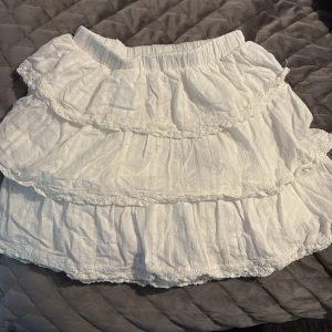 Vit volangkjol från Sky to Moon - Supersöt vit kortkjol från Sky to Moon med flera lager volanger och spetsdetaljer. Kjolen har resår i midjan och är tillverkad i ett lätt, luftigt tyg som passar perfekt till varmare dagar. Enkel att matcha med både toppar och sneakers.