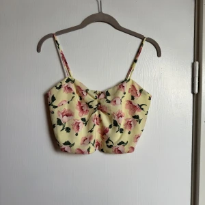 Gul blommig linnetopp med smock - Superfin ljusgul linnetopp med rosa blommor och gröna blad. Toppen har smala axelband, smockad rygg och en liten twist-detalj framtill. Croppad modell som är perfekt till höga jeans eller kjol. Materialet känns lätt och somrigt. Oanvänd 