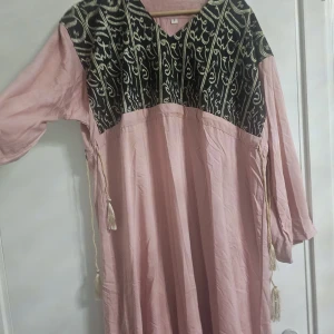 Lång rosa klänning med broderad överdel - Säljer en lång rosa klänning med svart och guld broderad överdel och detalj längst ner. Klänningen har v-ringning, långa ärmar och dekorativa snören med tofsar på sidorna. Materialet känns lätt och flowy, perfekt för dig som gillar unika detaljer.