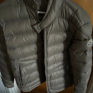 Beige Moncler dunjacka herr - Skick 9,5/10. Nypris 13000kr. Säljer en snygg beige dunjacka från Moncler med klassisk logga på ärmen. Jackan har hög krage, dragkedja och tryckknapp framtill samt två fickor med dragkedja. Perfekt för kyliga dagar och riktigt stilren look. 
