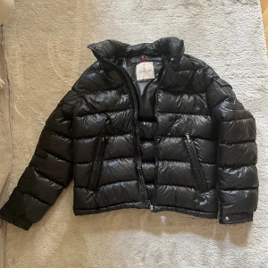 Svart pufferjacka från Moncler - Säljer en svart pufferjacka från Moncler med glansig finish och klassisk logga på ärmen. Jackan har hög krage, två dragkedjefickor framtill och snygga detaljer som tryckknappar vid ärmslut. Perfekt för kalla dagar när du vill hålla stilen. (Lappen på insidan har trillat bort, har intyg, garanterat äkta. Köpt på Moncler Birger Jarlsgatan Stockholm) 