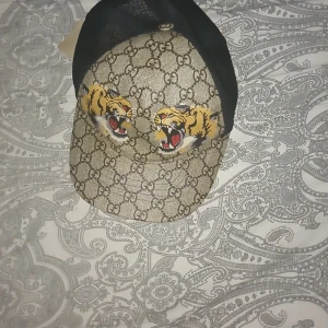 Gucci keps med tigertryck och mesh - Snygg keps från Gucci med klassiskt GG-mönster i beige och brunt, prydd med två tigrar framtill. Baksidan är i svart mesh och justerbar rem med gröna och röda detaljer. Perfekt statement-piece för dig som vill sticka ut.