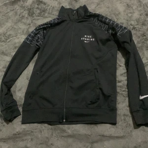 Nike Running svart vindjacka XL - Svart vindjacka från Nike Running i storlek XL. Jackan har hel dragkedja, höga muddar och grafiskt mönster i vitt på axlarna. Nike-logga och text på bröstet. Perfekt för löpning och träning, med sportig och clean look.