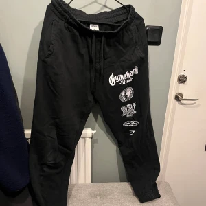 Svarta mjukisbyxor från Gymshark M - Svarta mjukisbyxor från Gymshark i storlek M med vita tryck och loggor på ena benet. Byxorna har resår i midjan, snörning och muddar vid bensluten. Perfekta för chill eller gymmet, riktigt sköna och snygga.