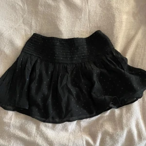 Svart kortkjol - Supersöt svart kortkjol från Gina Tricot med små glitter prickar, veckad midja. Kjolen är lätt utsvängd och har ett tunt, luftigt tyg som ger en flowy känsla. Perfekt att styla med en enkel topp eller stickad tröja. Har inte använt den som kjol då den är för liten men har använt den som topp några gånger!