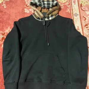 Svart burberry hoodie - Svart hoodie med stor ficka framtill och rutmönstrad huva i beige, svart och rött. Huvan har snörning med coola detaljer. Perfekt för dig som gillar en stilren look med en twist. Långärmad och skön passform.