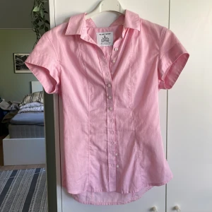 Rosa kortärmad skjorta från Shirt Factory - Snygg rosa skjorta från The Shirt Factory med klassisk krage och korta ärmar. Skjortan är figursydd med pärlemorknappar och är lättstruken. Perfekt för dig som gillar stilrena och färgglada plagg.