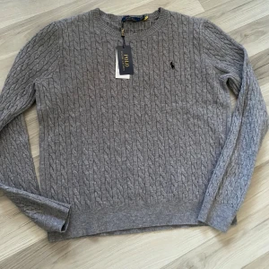 Grå kabelstickad tröja Polo Ralph Lauren - Kabelstickad grå tröja från Polo Ralph Lauren med rund halsringning och broderad logga på bröstet. Tröjan har lång ärm och ribbade muddar. Perfekt för dig som gillar klassisk och stilren look.