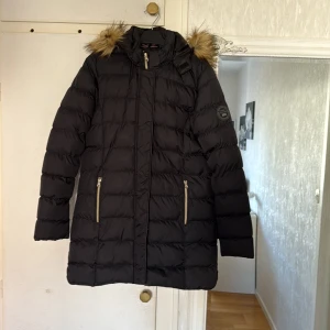 Svart lång pufferjacka Vinson Polo Club - Svart lång pufferjacka från Vinson Polo Club med huva och snygg fuskpäls. Jackan har guldiga dragkedjor, två fickor fram och quiltad design. Perfekt för kalla dagar med sin varma och stilrena look.