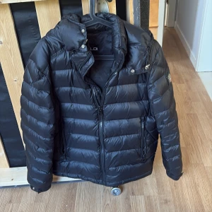 Svart pufferjacka från Bvaldi, M - Svart pufferjacka från Bvaldi med huva och dragkedja framtill. Jackan har ett stilrent guldigt logomärke på ärmen och två fickor med dragkedja. Perfekt för kalla dagar och riktigt snygg till streetwear-stilen.