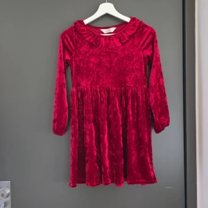 Röd sammetsklänning med volangkrage - Superfin röd sammetsklänning med lång ärm och volangkrage. Klänningen har en mjuk, glansig yta och ett klassiskt snitt med rynkad kjol. Perfekt för dig som vill sticka ut med en färgstark look.