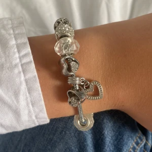 Silverfärgat armband med hjärtberlocker - Charmigt armband i silverfärg med flera glittriga och transparenta pärlor samt hjärtformade berlocker. Armbandet har detaljerade mönster och en öppen hjärtberlock som hänger. Perfekt accessoar för att lyfta din outfit. Köpt på en marknad utomlands.