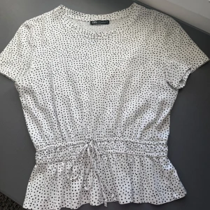 Prickig blus med knyt från Zara - Supersöt vit blus från Zara med svarta prickar, korta ärmar och rund hals. Midjan har smock och knytband som ger en snygg form och volangkant nedtill. Perfekt för dig som gillar lekfulla detaljer och en feminin look. Storlek M men skulle säga att den är liten i storleken och passar nån som har xs/s🥰