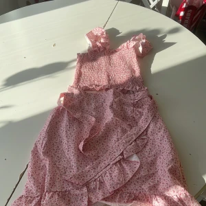 Rosa blommig volangklänning - Supersöt rosa klänning med småblommigt mönster och volangdetaljer. Smockad överdel med axelband och omlottkjol med volanger. Perfekt för sommarens alla tillfällen!