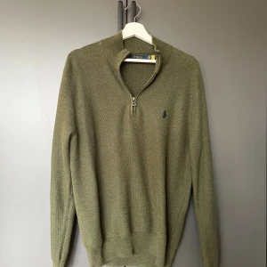 Olivgrön stickad tröja Polo Ralph Lauren - Snygg olivgrön stickad tröja från Polo Ralph Lauren med halv dragkedja och liten broderad logga på bröstet. Tröjan har lång ärm och ribbade muddar, perfekt för lager-på-lager under höst och vinter.