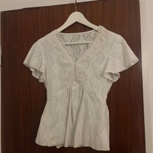 Vit blus med spets från SHEIN - Söt vit blus från SHEIN med broderade vågmönster och v-ringning dekorerad med spets. Blusen har korta, vida ärmar och är tillverkad i ett lätt material med hålbroderier som ger en luftig känsla. Perfekt för en fräsch och somrig look.