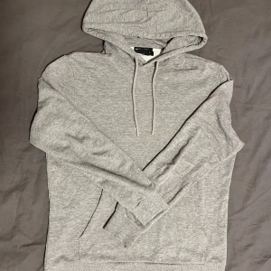 Grå hoodie  - Snygg grå hoodie med klassisk huva och snörning. Tröjan har långa ärmar, ribbade muddar och en stor magficka. Perfekt för chill dagar eller när du vill ha en enkel och stilren look.