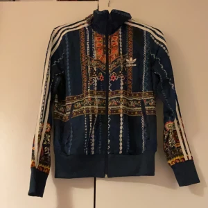 Mönstrad Adidas hoodie med dragkedja - Säljer en unik hoodie från Adidas med färgstarkt mönster i blått, rött och gult. Klassiska vita ränder längs ärmarna, dragkedja framtill och hög krage. Perfekt statement-plagg för dig som vill sticka ut.