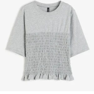 Grå topp med smock och volang - Grå topp från Divided med smockad front och volangkant nedtill. T-shirten har rund halsringning och korta ärmar. Perfekt för en chill och trendig look.