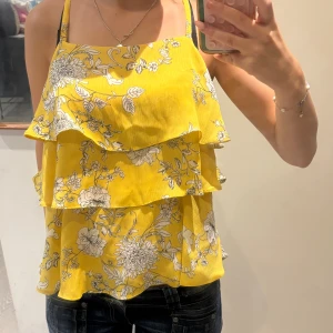 Gul blommig volangtopp River Island - Superfin gul linnetopp från River Island med vita blommor och tunna axelband. Toppen har flera lager volanger som ger en flowy och söt look. 