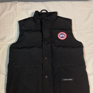 Svart dunväst från Canada Goose - (Säljer och byter mot en size S)Svart dunväst från Canada Goose med hög krage, tryckknappar och två stora fickor framtill. Klassisk logga på bröstet och diskret Canada Goose-märke på ena fickan. Perfekt för lager-på-lager och riktigt snygg till streetwear.