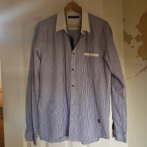 Randig skjorta från Selected Homme - Snygg skjorta från Selected Homme med blå och vita ränder, kontrasterande vit krage och bröstficka med vit kant. Klassisk passform och långa ärmar, perfekt för en stilren look. Skjortan har knäppning framtill och är tillverkad i mjuk bomull.